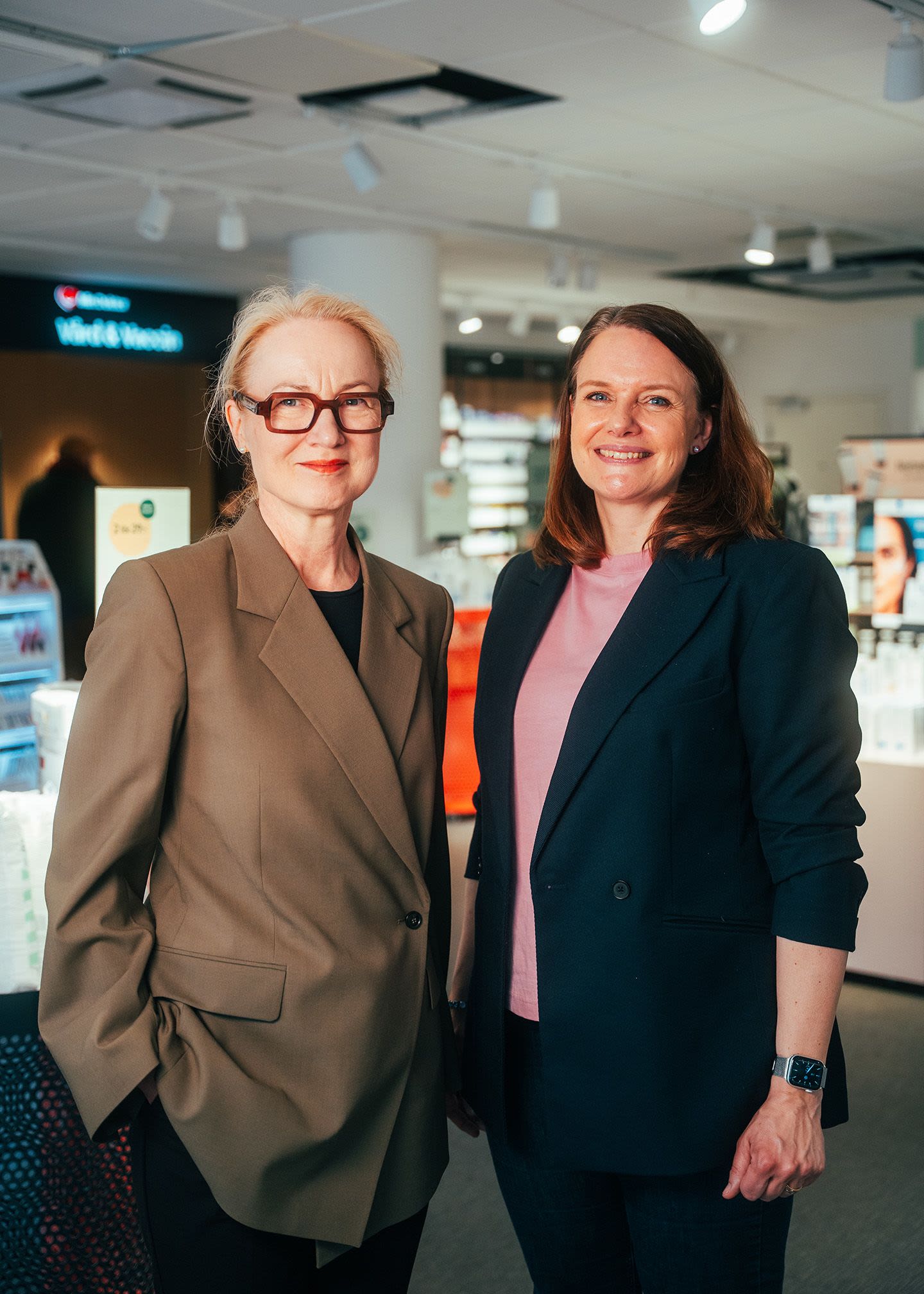 Two women in professional attire stand together smiling inside a pharmacy.
