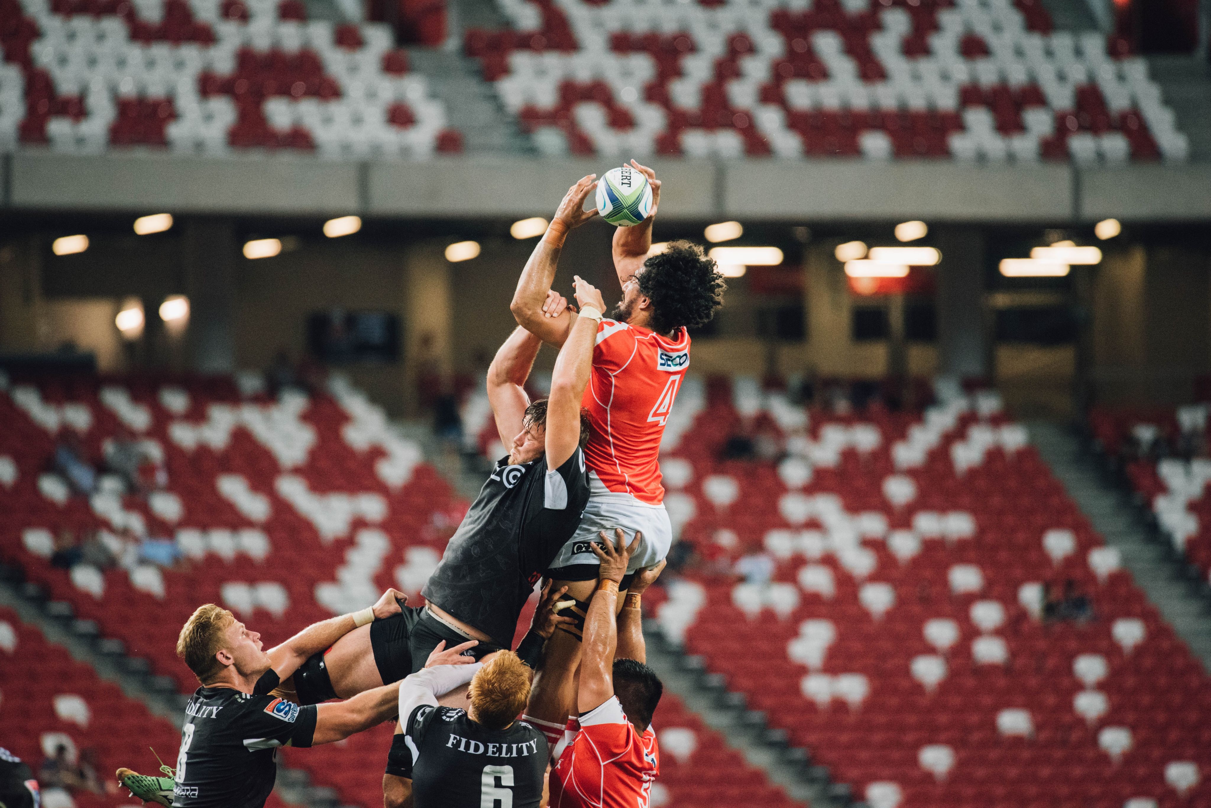 Two people playing sport and leaping to catch a rugby ball. One wears a red shirt and one wears a black shirt.