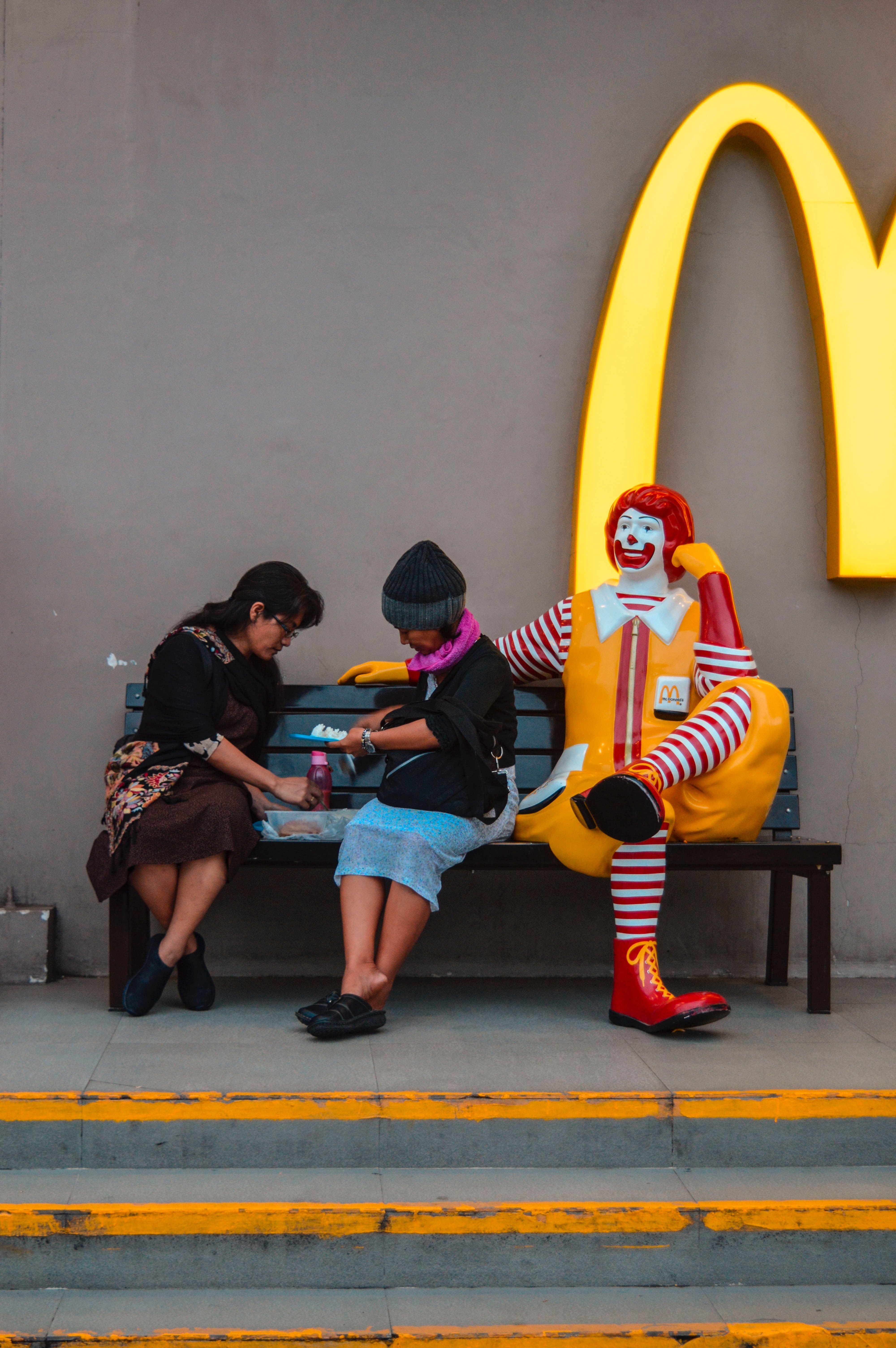 Two women sit at a McDonald's bench next to a Ronald McDonald statue.