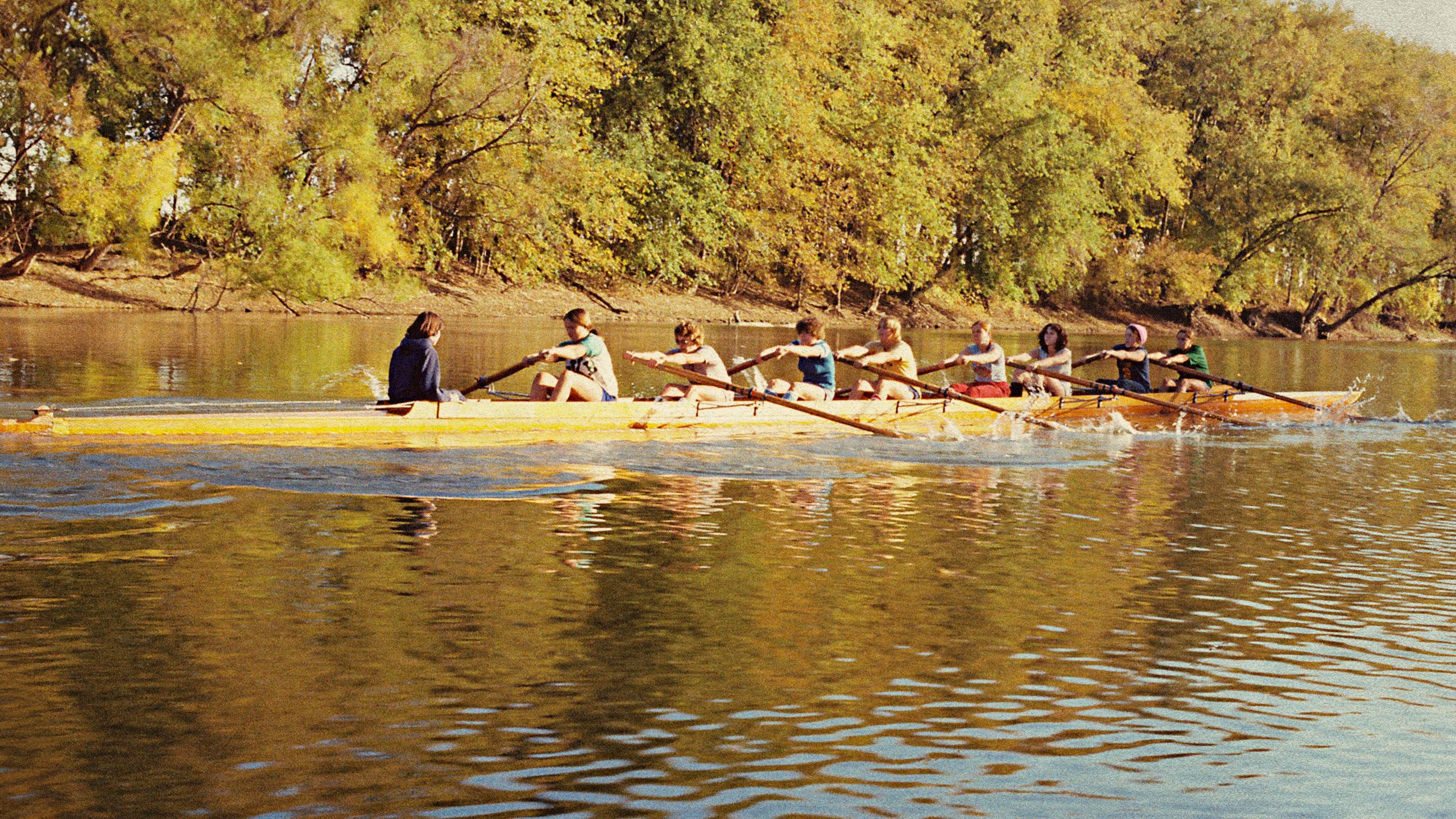 A rowing team paddles a long boat on a flat body of water in front of a forest.
