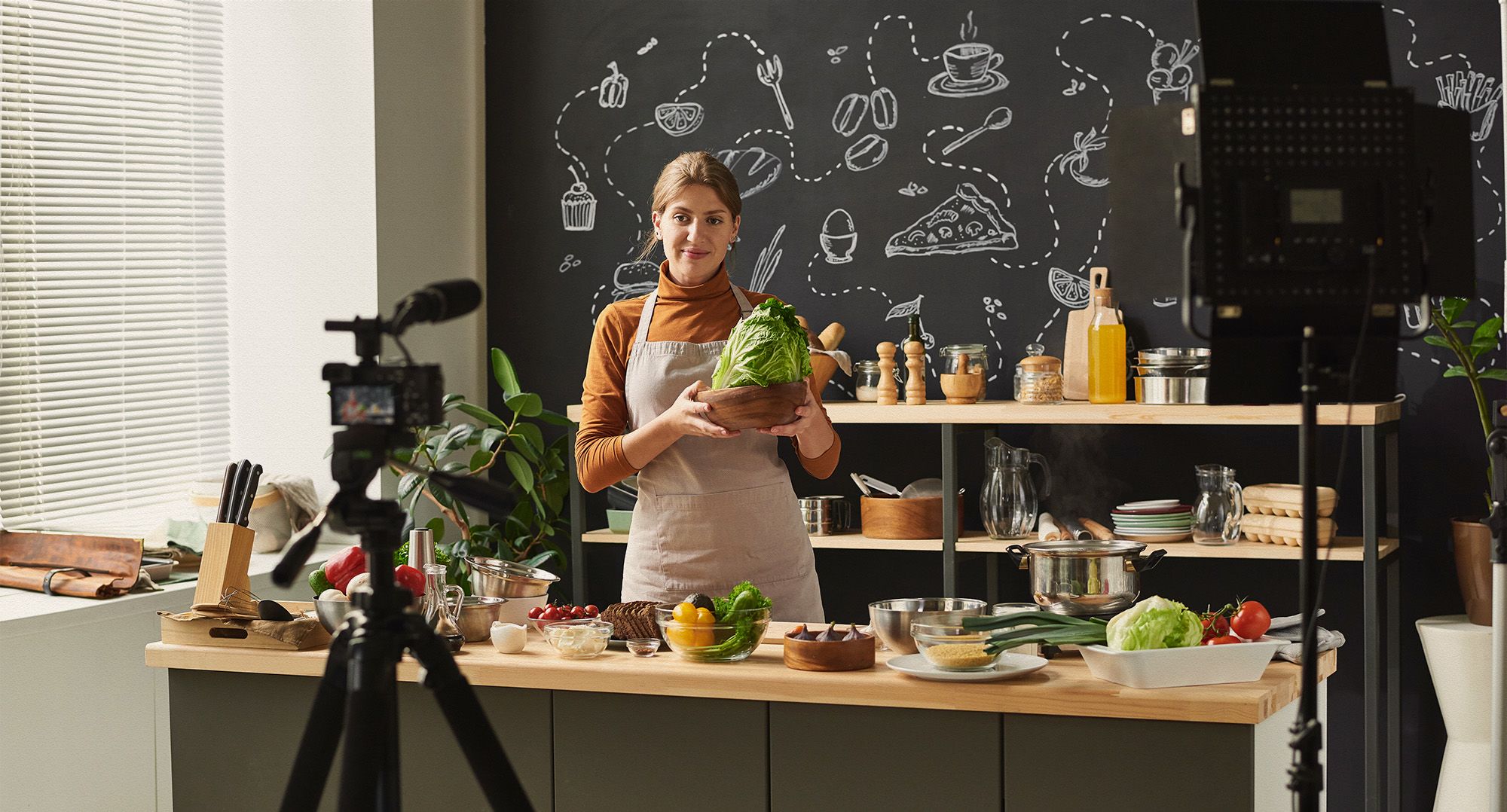 A smiling person wearing an apron and holding vegetables in a kitchen with a film camera in front of them.