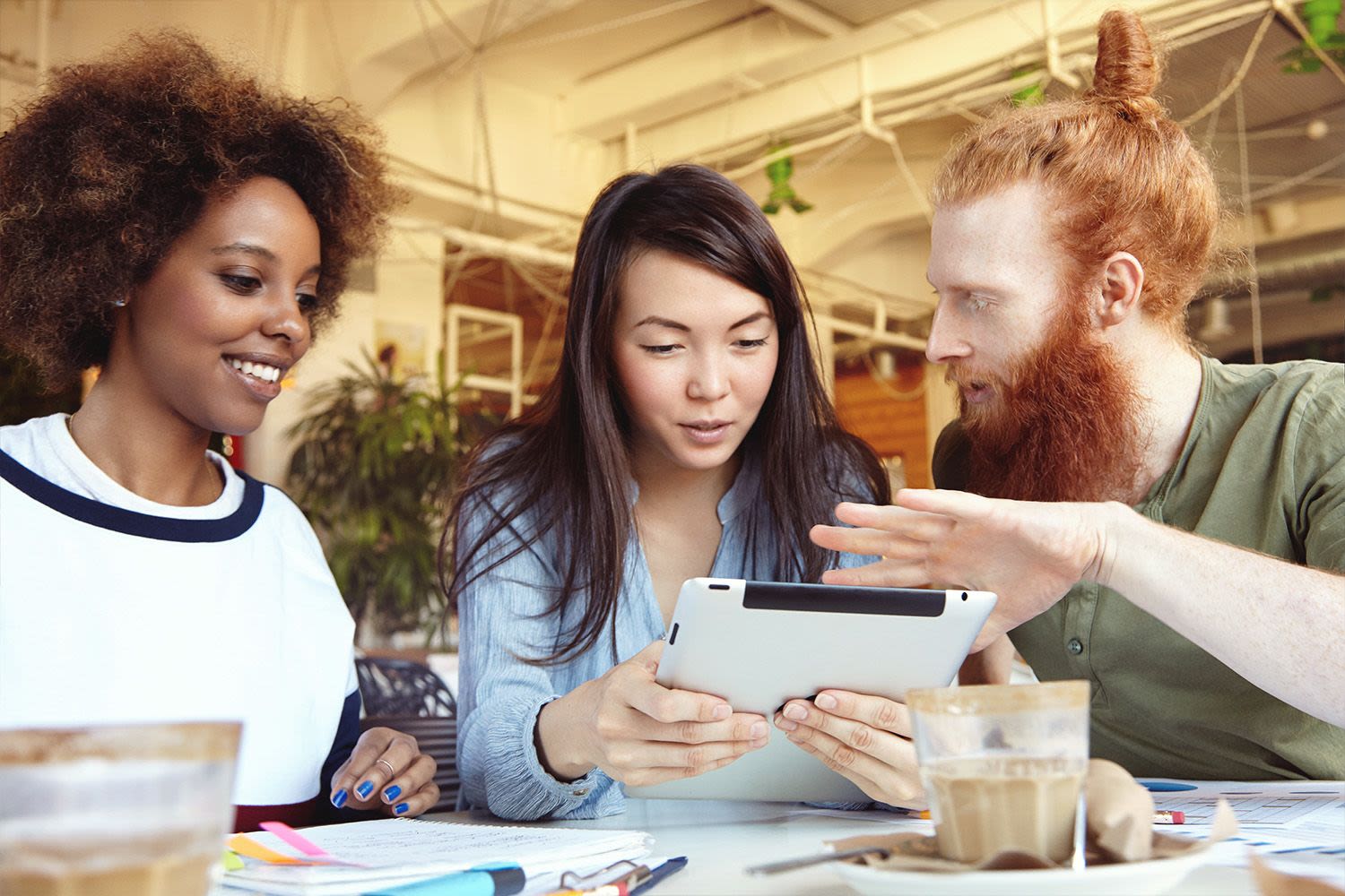 A woman with dark skin and black hair, a woman with light skin and black hair, and a man with white skin and red hair, sit working together in an office and looking at a tablet screen.