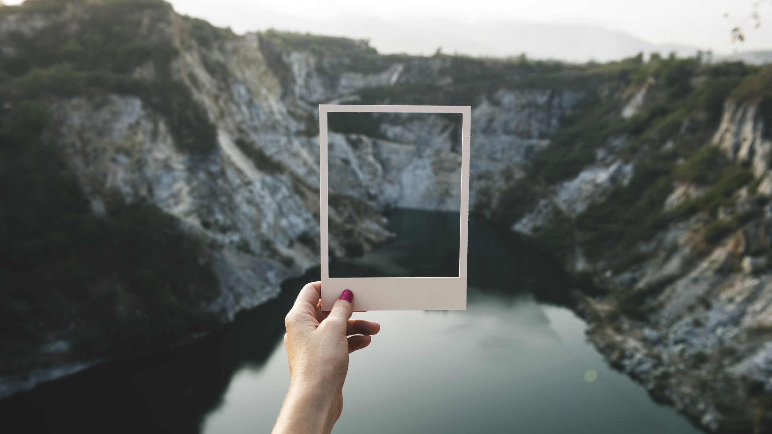A hand holding an empty polaroid frame in front of mountain and lake 