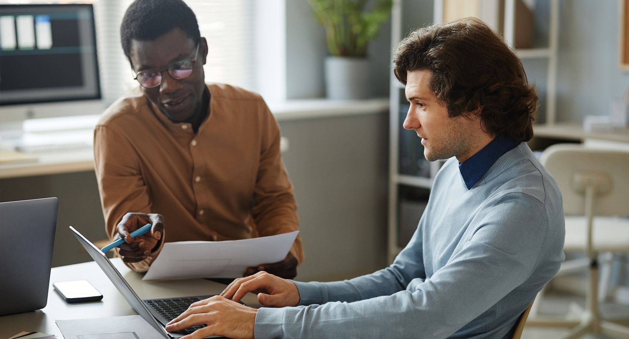 Two people, one in a blue sweater and one in a mustard button-down sit in front of one laptop, clearly in friendly discussion