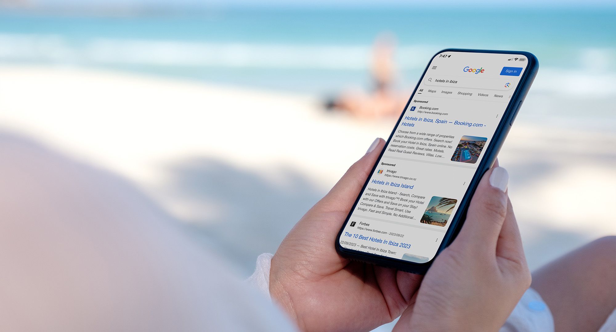 A phone displaying goodle search results for 'hotels in Ibiza', held by someone sitting on a beach