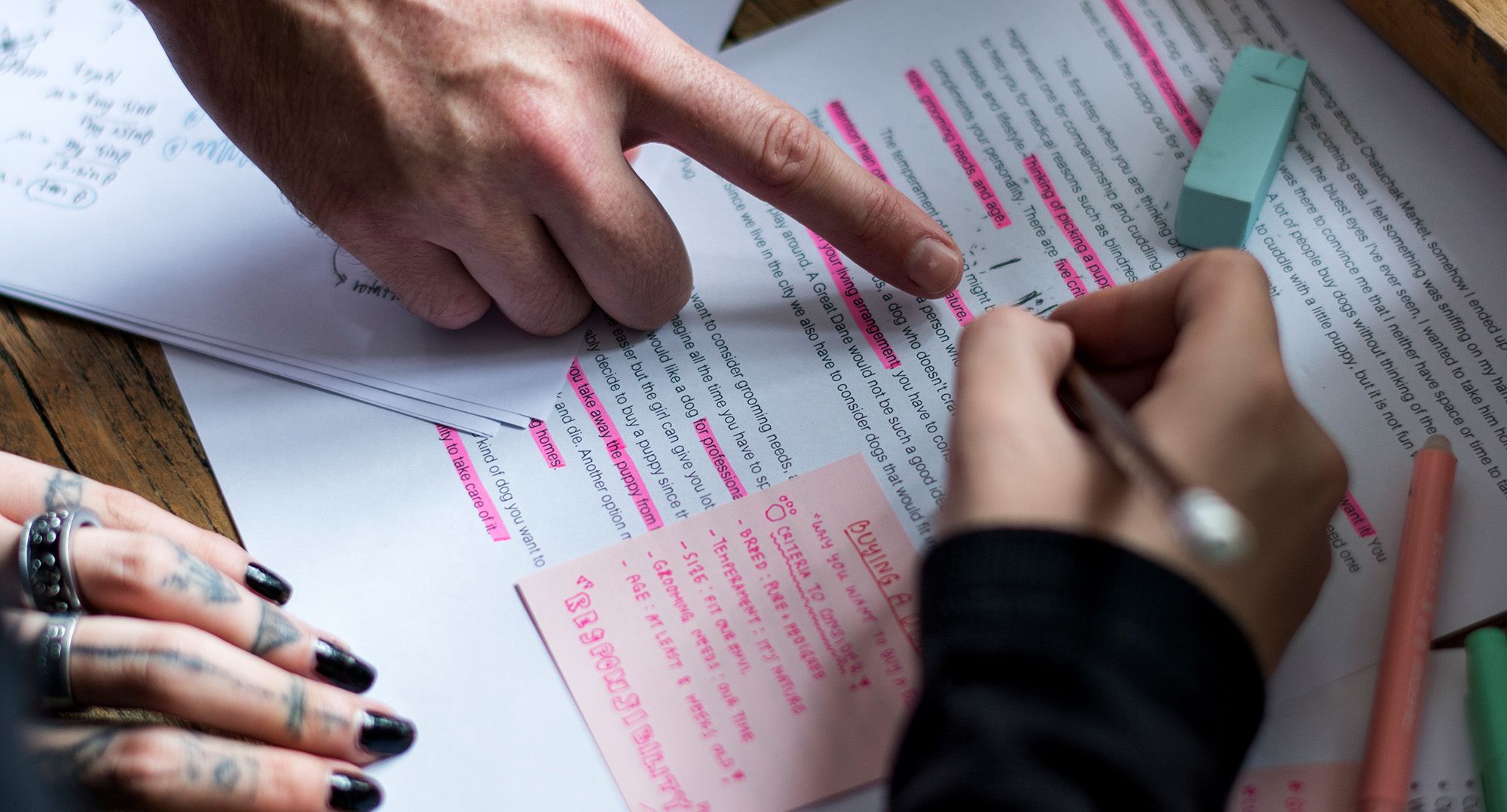 A page highlighted in pink, with a pink sticky note, pens scattered around, and three different people's hands pointing things out