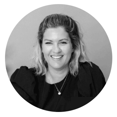 A black and white portrait in a circular frame of a person with shoulder-length wavy light hair, dark eyes, and a bright smile.