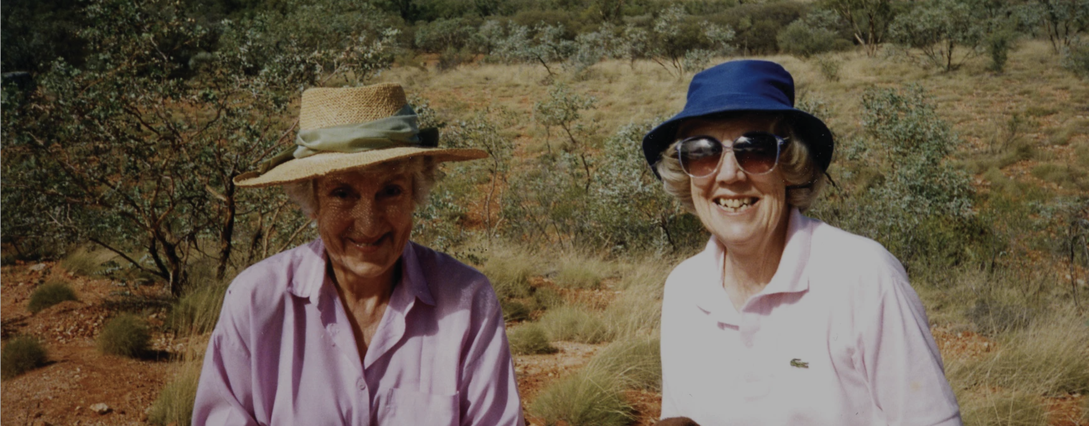 Two elderly women with white skin, wearing sunhats and sunglasses, stand together smiling.
