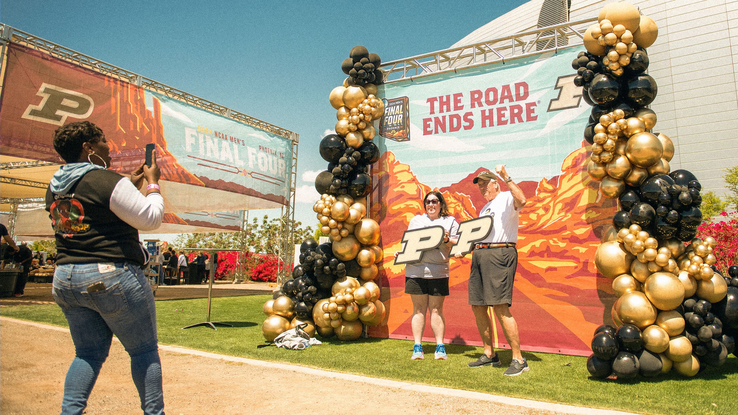 Two people wave while a woman takes a photo with a smartphone.