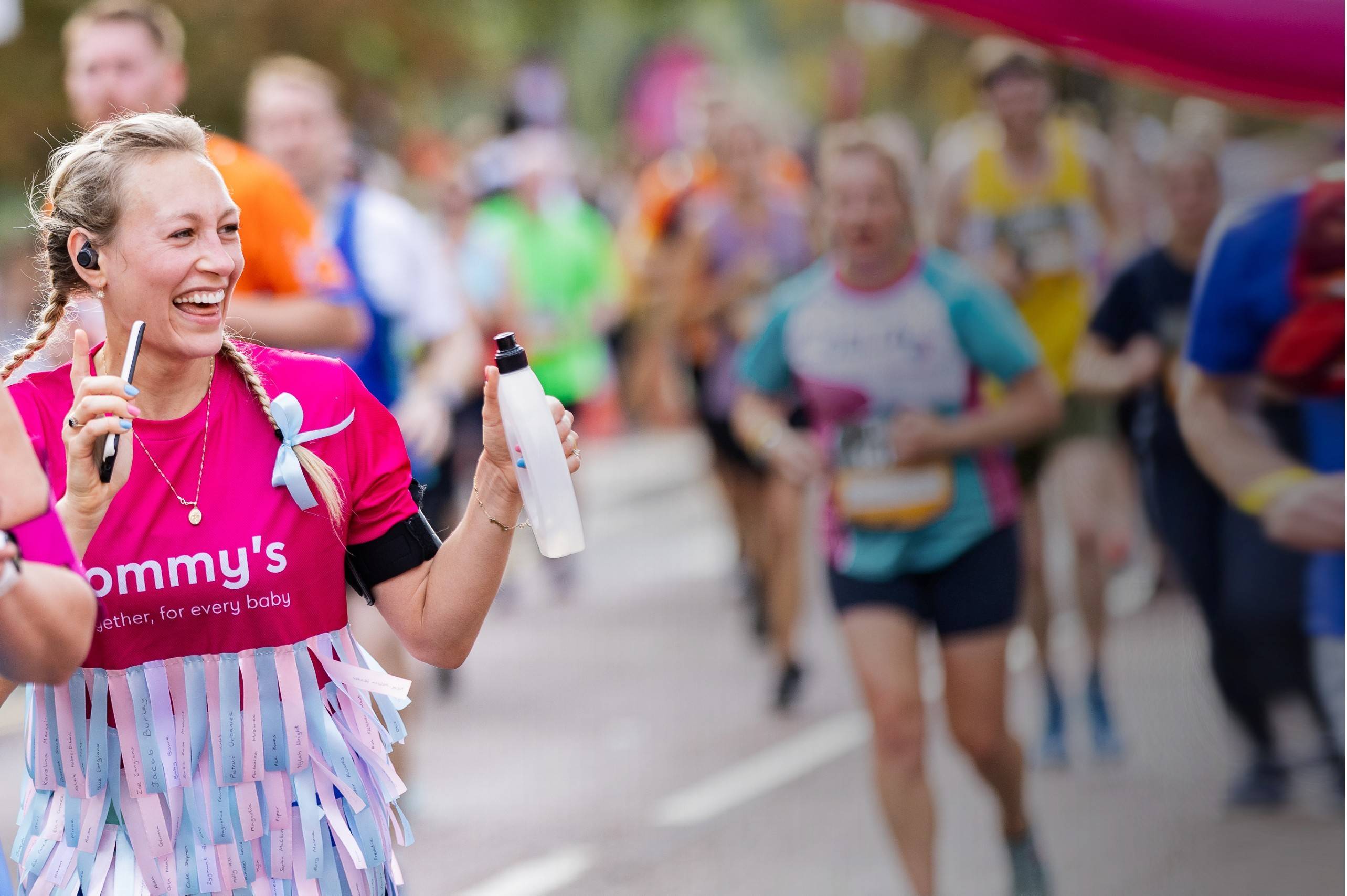 A woman with white skin and blonde plaited hair hods a drink bottle and smartphone, standing in front of joggers in the background.