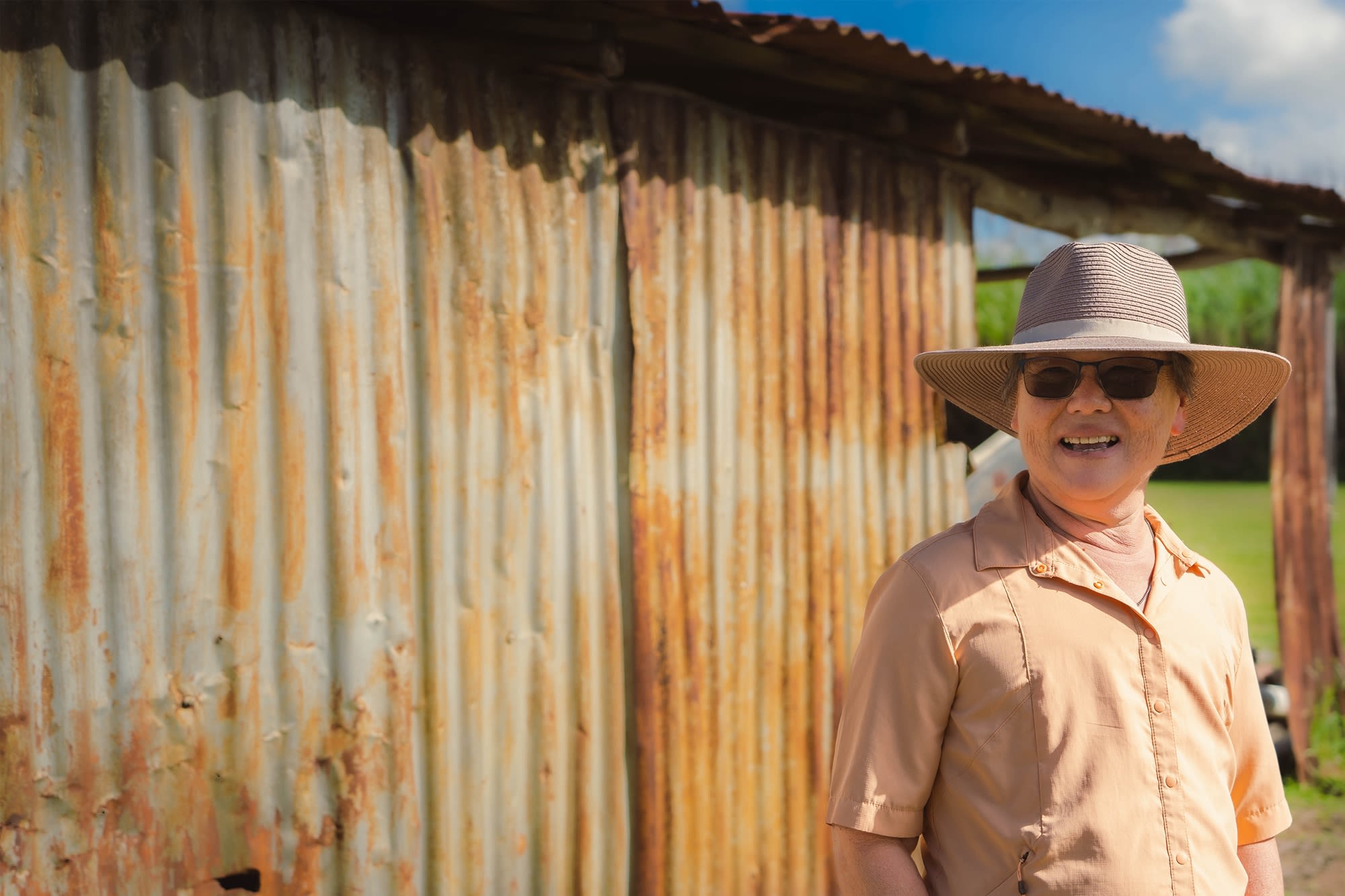 A woman with light skin wearing a cowboy had and light shirt stands in front of a rusty shed.