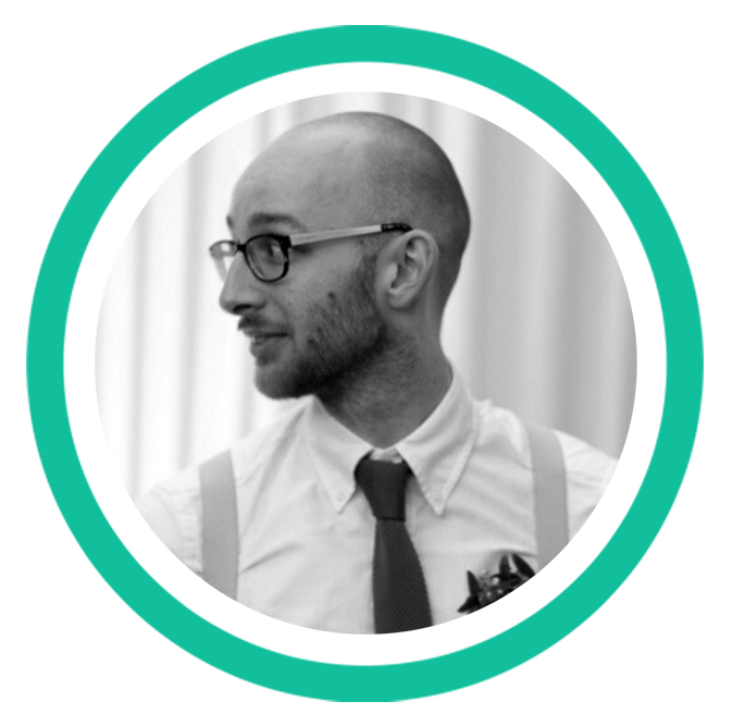 A black and white headshot of a person with a shaved head, a short beard, and dark rimmed glasses. 