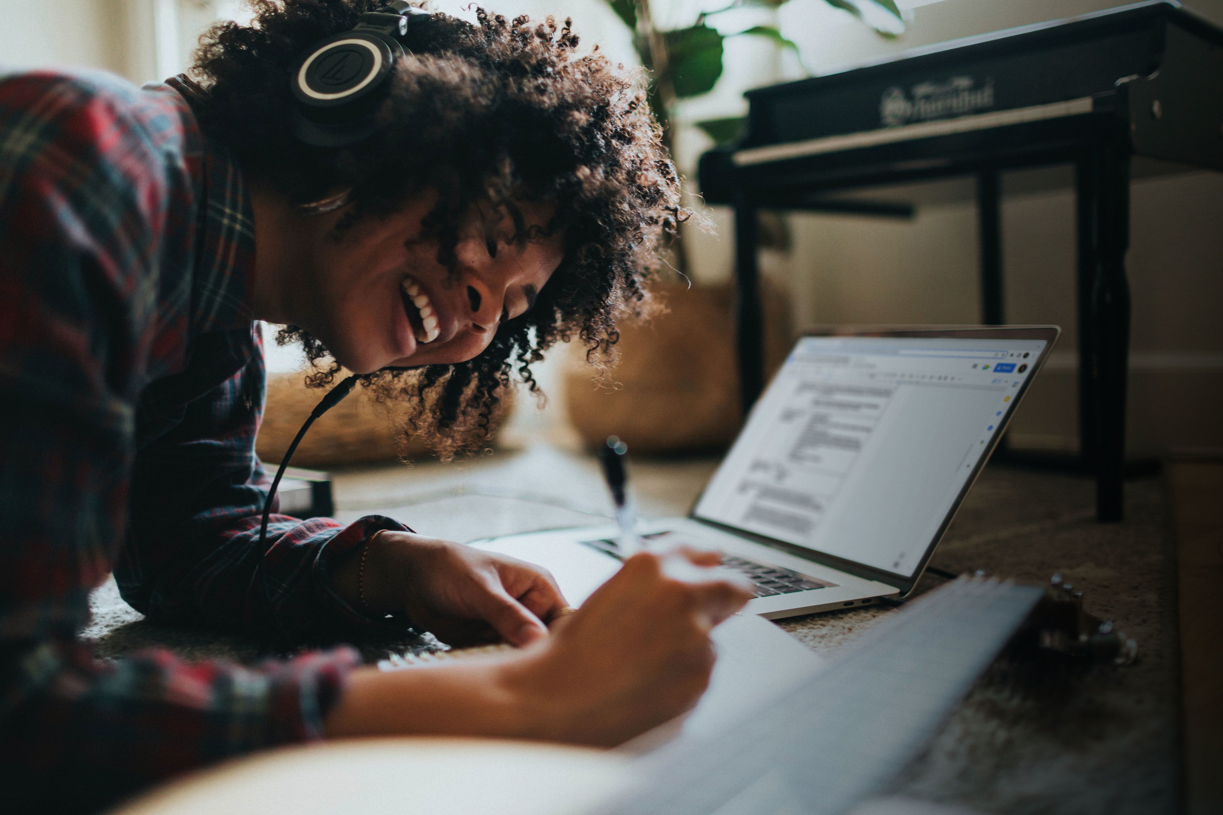 A smiling person working on a laptop, they're also writing with a pen in a notebook.