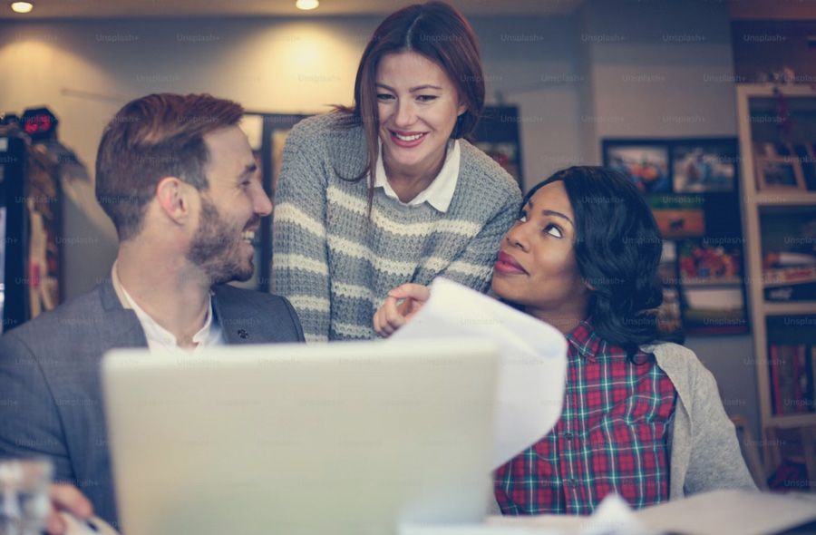 three people of different genders and ethnicities gather, smiling, around a laptop