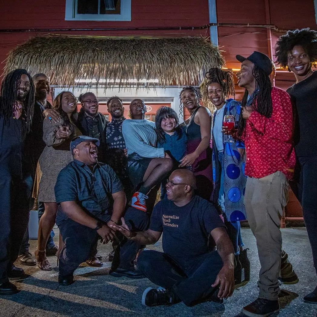 A large group of African American hospitality workers arranged in a loose curve. Two older people sit in front of them. All are smiling or laughing with each other. The person second from the far right wears a bright red shirt and holds a wine glass filled with something in a matching colour. There is a dried front arrangement that hangs over the open door behind them