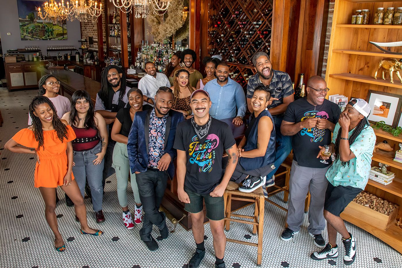 As earlier, seventeen young African Americans gather around chairs and tables, beaming and facing the camera. Several wear Turning Tables t-shirts, which are black with stylised text in gold, pink and light blue. Because the image is square-edged, it is clear they are in a bar, with a wine rack in the middle background, and a bar behind on the left. To the right is a light pine shelf filled with sculptures, books and framed art. The floor is black and white mosaic tile