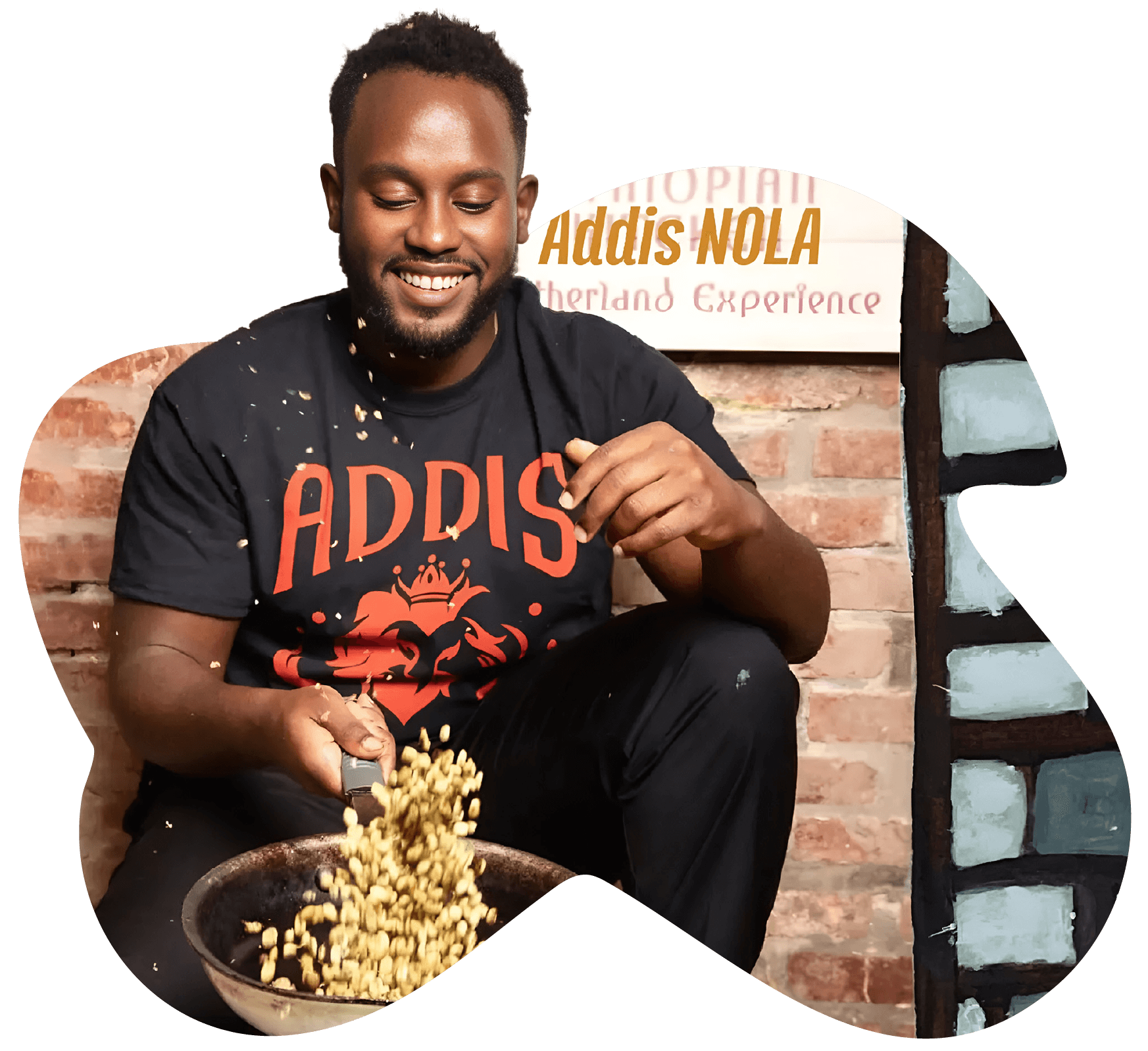 A smiling young Black person tosses peanuts in a pan. They have short hair and look away from the camera at the frying pan. They are wearing black trackpants and a black t-shirt with a red "ADDIS" logo and a graphic of a red stylised lion. In the background is a brick wall and a sign saying "Addis NOLA Motherland Experience"