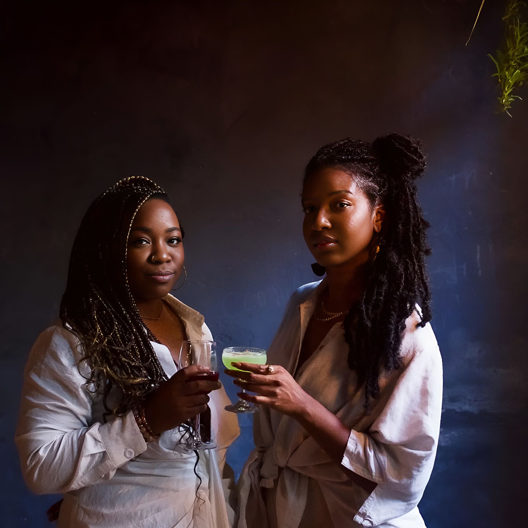 Two young Black people with long, braided hair stand slightly apart from each other with angled shoulders, gazing into the camera. They smile slightly and hold their drinks up. The person on the left holds a glass of red wine, the person on the right holds a lime green cocktail. Both are wearing bone-coloured linen jackets. The background is dark slate grey