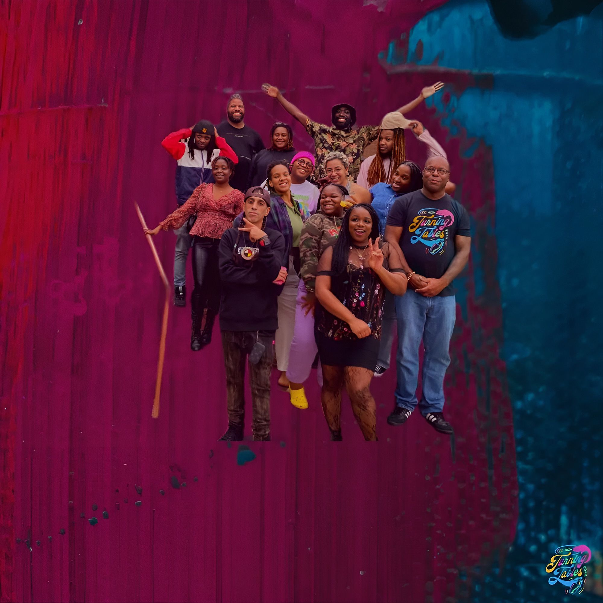 Fourteen people, all people of colour, stand in a group in front of a background that is three quarters magenta, one quarter dark turquoise. They all smile forward; two flash peace signs, two adjust their caps, and one has their hands joyfully held above their head. They are all dressed in casual, cheerful colours, and a bald person at the front right wears a Turning Tables logo t-shirt