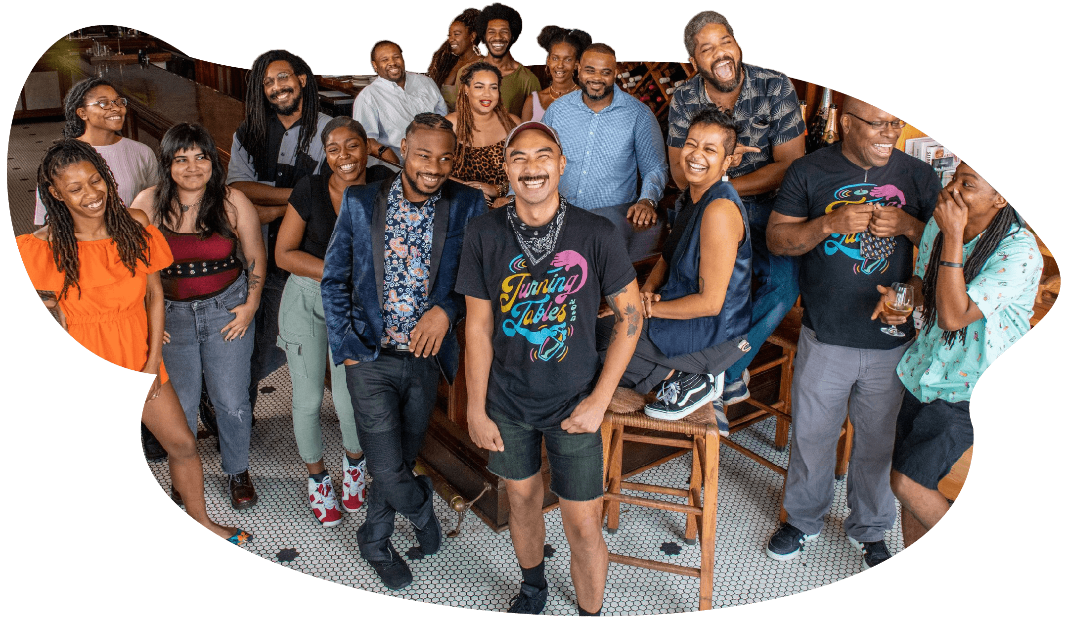 Seventeen young African Americans gather around chairs and tables, beaming and facing the camera. Several wear Turning Tables t-shirts, which are black with stylised text in gold, pink and light blue. They are dressed casually and standing casually. The floor is black and white mosaic tile