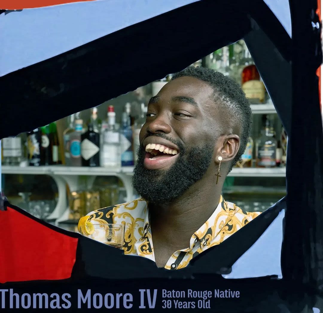 Headshot of a Black person with short hair who laughs at something to the left of focus. They have a short beard, a dangling golden crucifix earring in in their right ear, and wear a gold, black and white batik collared shirt. They stand in front of two shelves of spirits. The edges of the image have thick drawn angular bars of black and red and pale blue framing their face.
