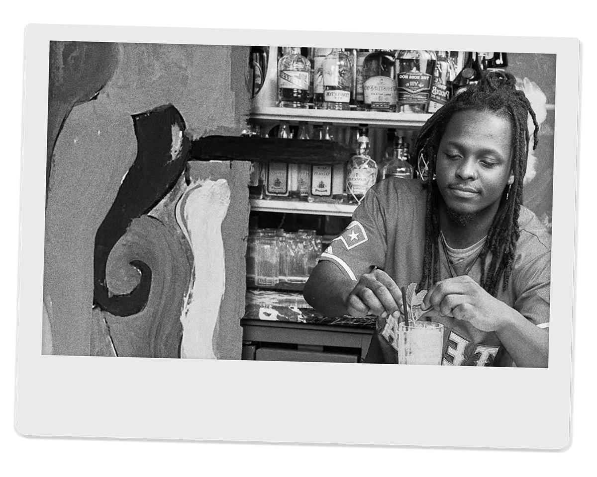 Black and white photo of a young African American mixes a drink in front of a cabinet of spirit bottles. They stand to the right of frame, looking down at the tall clear glass they garnish with small leaves. To the left is a wall painted in large swirls
