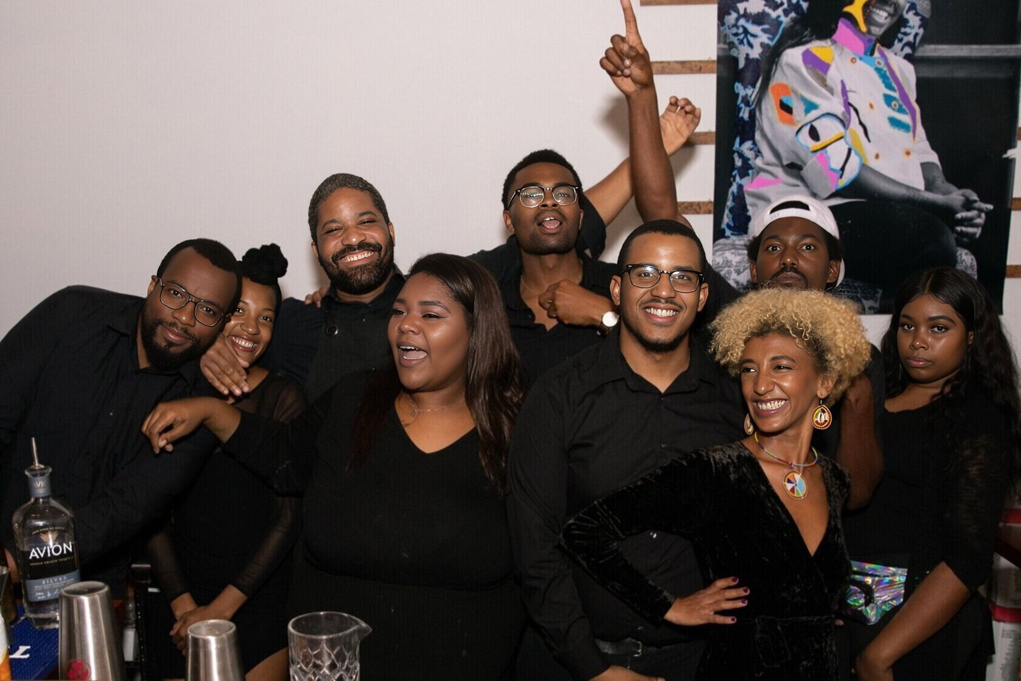 Nine African American people in black clothes gather in front of a white wall. In the right corner, half of an artwork is visible, showing the bottom half of a woman sitting down. The people are mid-conversation, laughing and with their arms around each others' shoulders