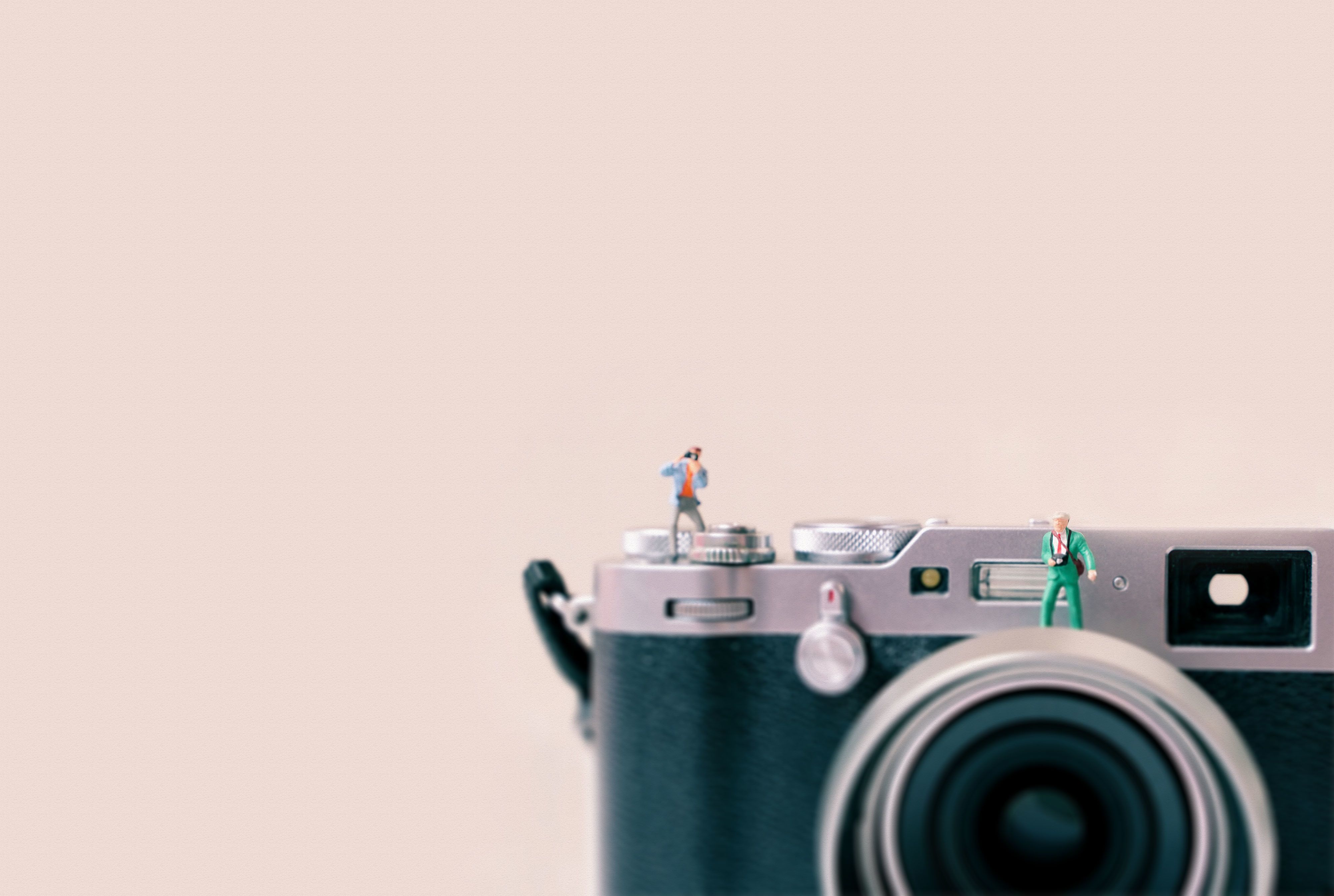 A film camera with tiny model people standing on top of it. The camera is set against a pastel pink background.