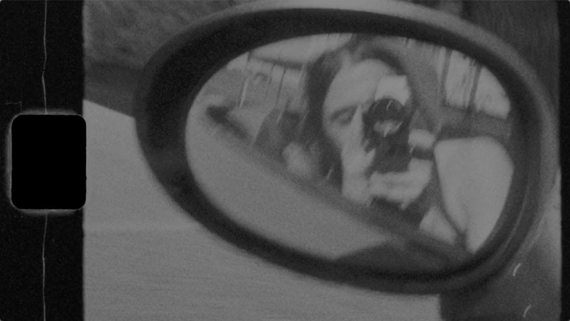 black and white image of car review mirror with a reflection of a woman filming with a super 8 camera