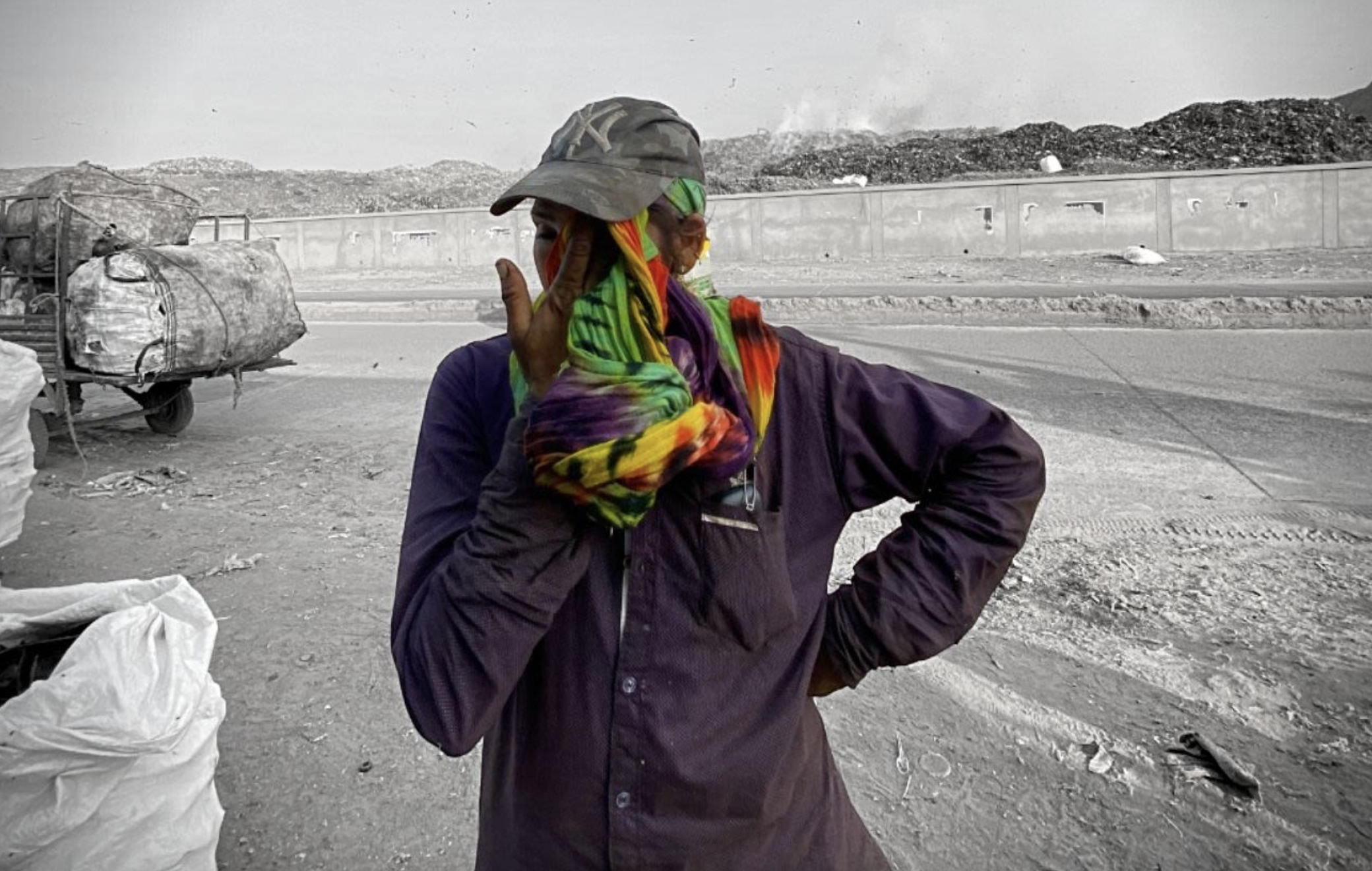 A dark-skinned person wearing a black cap and purple boiler suit wipes their eyes with a colourful rainbow kerchief. They're standing in front of a cart carrying large sacks and, further away, a wall. The background is in greyscale.