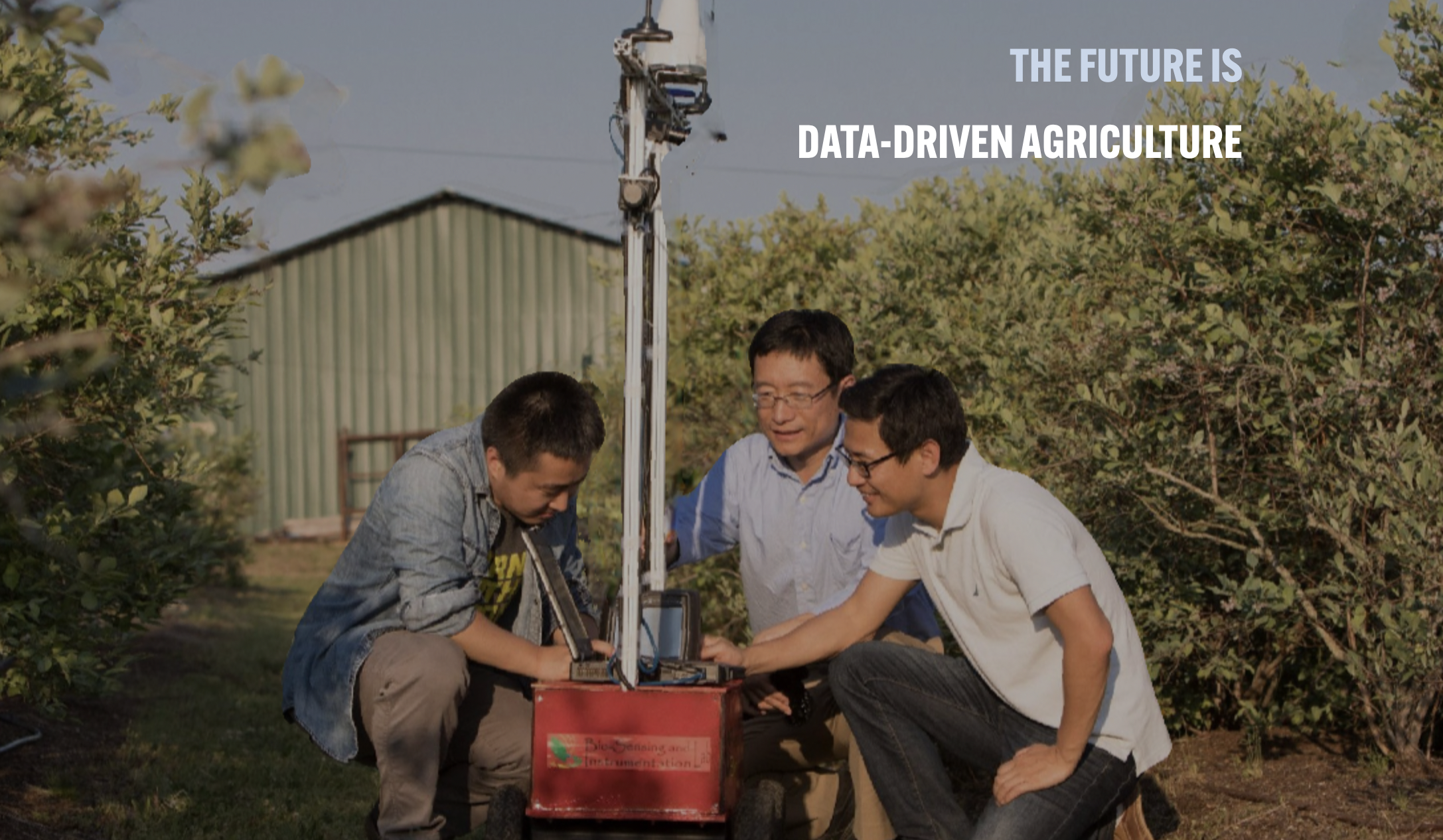 Three researchers crouch around a tall piece of robotic equipment that is perched on top of a red crate. They are outside a green corrugated iron shed and several dusty green bushes.