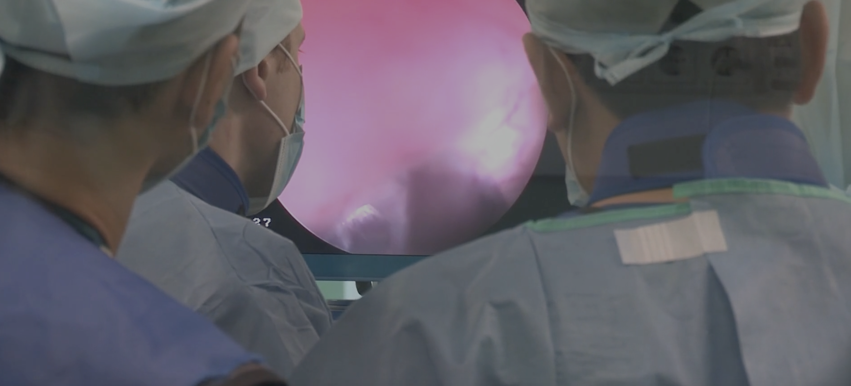 Three researchers wearing blue medical scrubs face away from the camera, looking toward an oval screen or window filled with purple light.