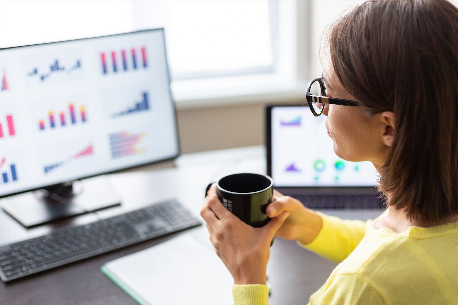 A person with long dark hair in a yellow top drinking coffee while working at a computer.